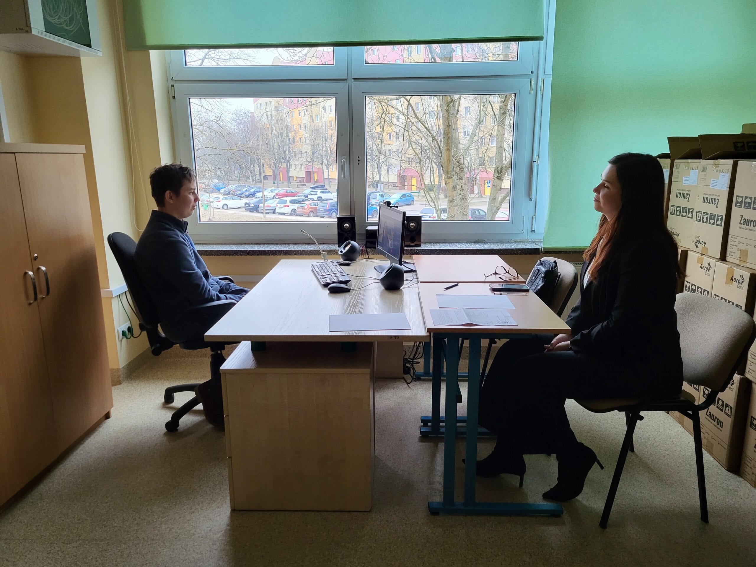 Dwie osoby siedzą naprzeciw siebie przy biurku w szkolnym gabinecie, prowadząc rozmowę przy komputerze ustawionym przy dużym oknie z widokiem na osiedle i drzewa.