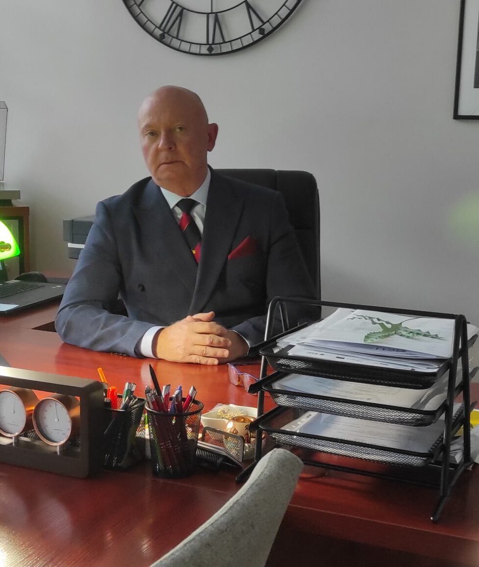 Osoba siedząca za biurkiem w eleganckim gabinecie. Ma na sobie ciemny garnitur z krawatem i poszetką. Na biurku znajdują się tacki na dokumenty, przybory do pisania, zegar biurkowy oraz sprzęt biurowy. W tle widać fragment ściany z dużym dekoracyjnym zegarem.