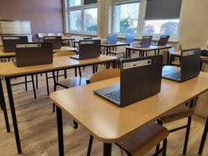 Sala lekcyjna z ustawionymi w rzędach drewnianymi ławkami, na których stoją otwarte laptopy z naklejkami informującymi o finansowaniu z programów rządowych i unijnych. W tle widoczne są duże okna oraz zaciemniona roleta.