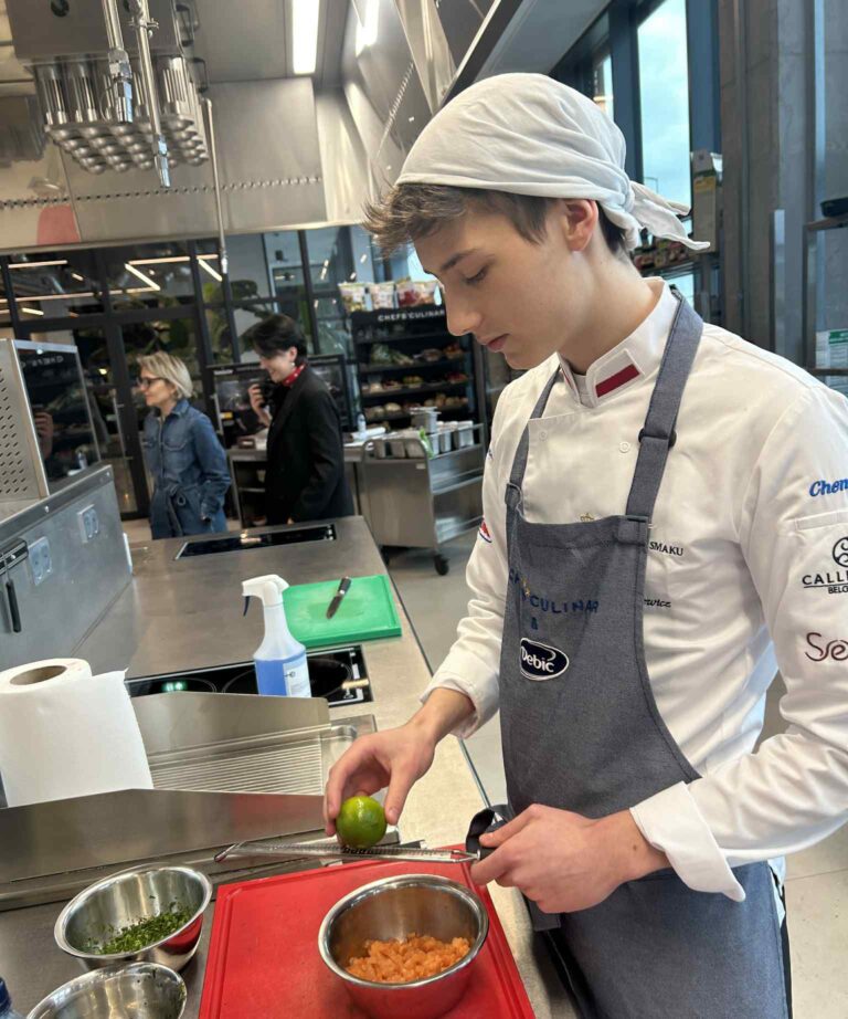 Osoba w stroju kucharskim pracuje w profesjonalnej kuchni. Stoi przy blacie, trzymając limonkę nad metalową miską z przygotowywanym składnikiem. Obok znajdują się miski z posiekanymi dodatkami oraz deska do krojenia. W tle widoczne są stanowiska kuchenne i kilka osób obserwujących pracę w kuchni.