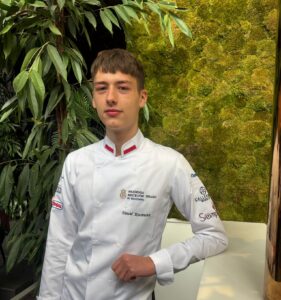 Uczeń w białym stroju kucharskim stoi przy ladzie na tle zielonej, roślinnej ściany dekoracyjnej.