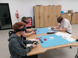 rzech uczniów siedzi przy dużym stole w pracowni technicznej i korzysta z kontrolerów oraz gogli FPV do nauki obsługi dronów. Na blacie leżą przewody, narzędzia i materiały dydaktyczne. W tle znajdują się wysokie drewniane szafki oraz stanowisko warsztatowe
