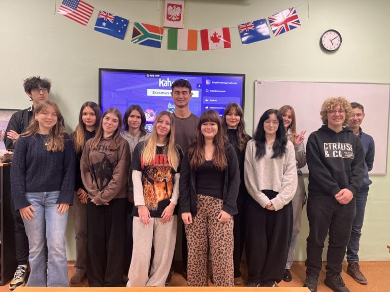 Grupa uczniów stoi w sali lekcyjnej przed ekranem multimedialnym, na którym wyświetlany jest quiz Kahoot związany z projektem Erasmus+, a nad nimi widoczne są flagi różnych państw.