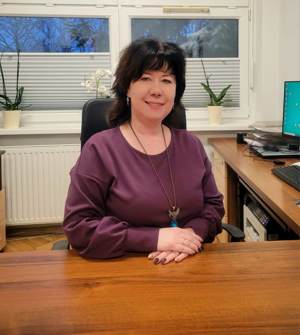 Osoba siedząca za biurkiem w gabinecie. Ma na sobie fioletową bluzę i naszyjnik, a dłonie trzyma złożone na blacie. W tle widać okno z roletami, dwie doniczki z roślinami oraz stanowisko komputerowe.