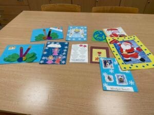 Na drewnianym stole leży dziewięć kolorowych kartek świątecznych wykonanych ręcznie. Kartki przedstawiają różne motywy, m.in. Mikołaja, aniołki, choinki, śnieżynki i ozdoby. W tle widać fragment krzeseł i szafek