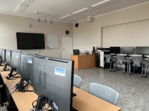 Sala komputerowa z rzędem monitorów ustawionych na długim biurku po lewej stronie. W głębi pomieszczenia znajdują się biurko nauczyciela, tablica, duży monitor ścienny oraz dodatkowe stanowiska komputerowe z szarymi krzesłami
