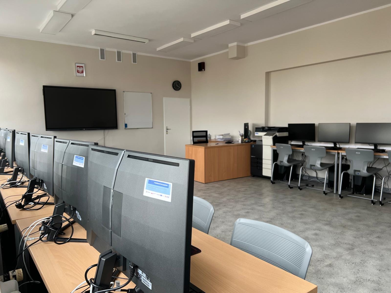 Sala komputerowa z rzędem monitorów ustawionych na długim biurku po lewej stronie. W głębi pomieszczenia znajdują się biurko nauczyciela, tablica, duży monitor ścienny oraz dodatkowe stanowiska komputerowe z szarymi krzesłami