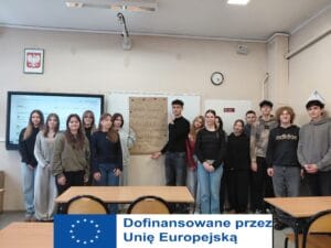 Grupa młodzieży pozuje do zdjęcia w sali lekcyjnej. W centrum dwoje uczniów trzyma arkusz szarego papieru z ręcznie wypisanymi zasadami ekologicznymi w języku angielskim i polskim (m.in. Rethink, Refuse, Reduce, Reuse). Po lewej stronie widoczny jest interaktywny monitor z wyświetloną stroną internetową, a nad nim godło Polski. Uczniowie stoją w swobodnych strojach, a w tle widać zegar ścienny oraz białą tablicę.