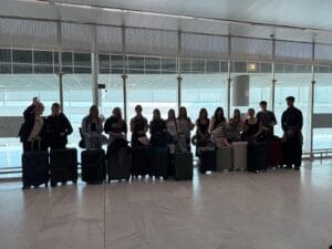 Grupa młodych osób pozuje do zdjęcia w jasnej hali lotniskowej. Stoją w rzędzie, trzymając przed sobą walizki podróżne, niektórzy machają w stronę aparatu