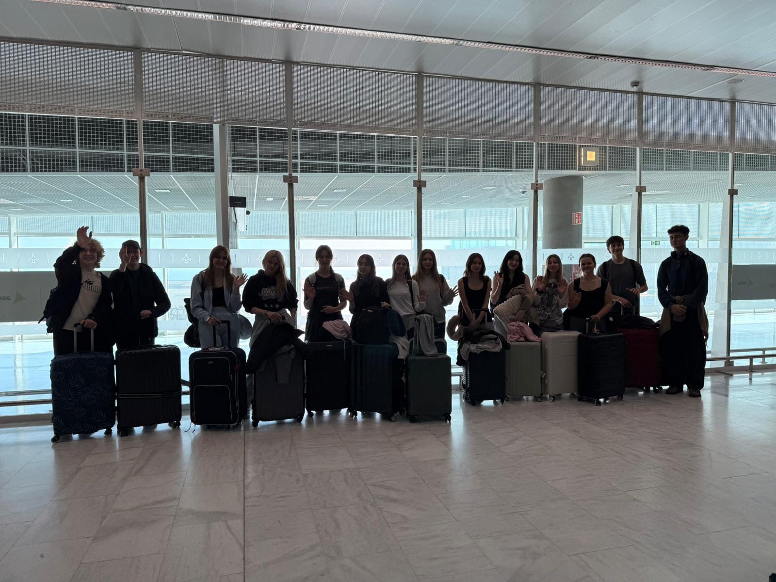 Grupa młodych osób pozuje do zdjęcia w jasnej hali lotniskowej. Stoją w rzędzie, trzymając przed sobą walizki podróżne, niektórzy machają w stronę aparatu