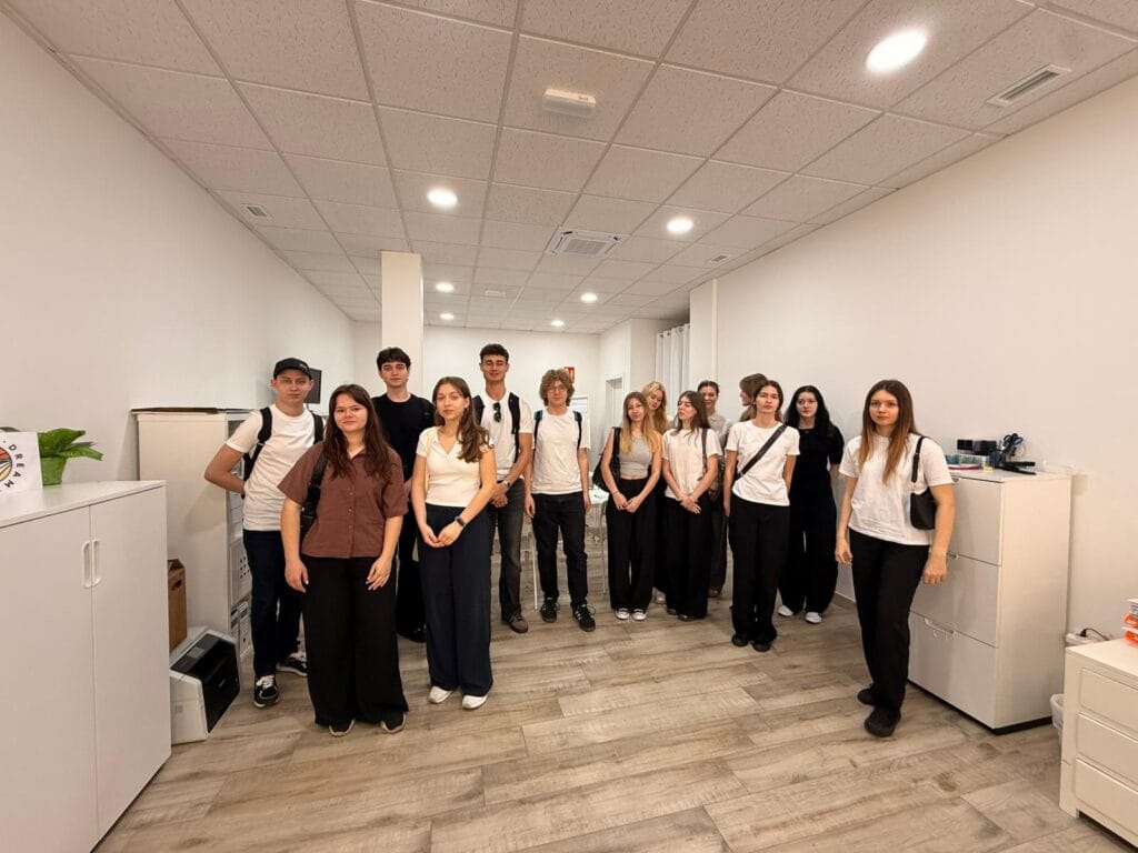 Grupa młodzieży stoi w jasnym, nowoczesnym pomieszczeniu biurowym lub szkolnym o białych ścianach i jasnej podłodze z paneli. Większość osób ubrana jest w białe koszulki i ciemne spodnie. Wzdłuż ścian stoją białe szafki biurowe, a na suficie zamontowane są kasetonowe panele z punktowym oświetleniem LED