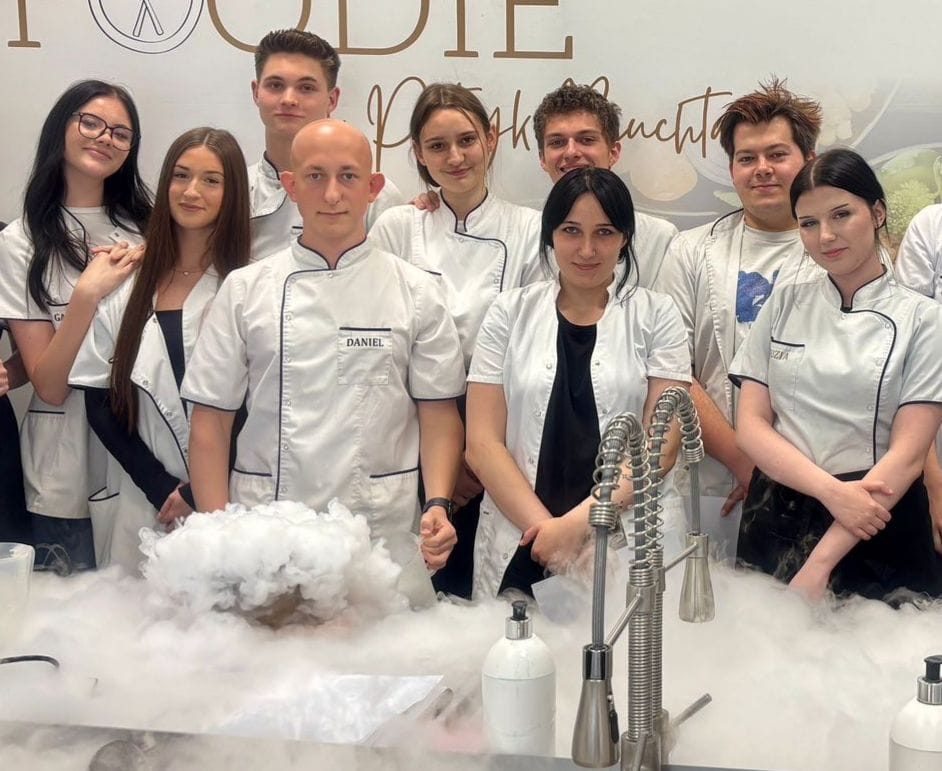 Zbliżenie na grupę młodych kucharzy pozujących w kuchni warsztatowej. W centralnym punkcie kucharz o imieniu Daniel stoi za blatem, z którego unoszą się kłęby białej pary z suchego lodu. Obok niego oraz w tle stoją pozostali uczestnicy szkolenia w białych kitlach. Wszyscy patrzą w stronę aparatu. W górnej części kadru widoczny jest fragment napisu „FOODIE by Patryk”. Na pierwszym planie znajduje się profesjonalna bateria kuchenna z elastyczną wylewką oraz białe dozowniki.