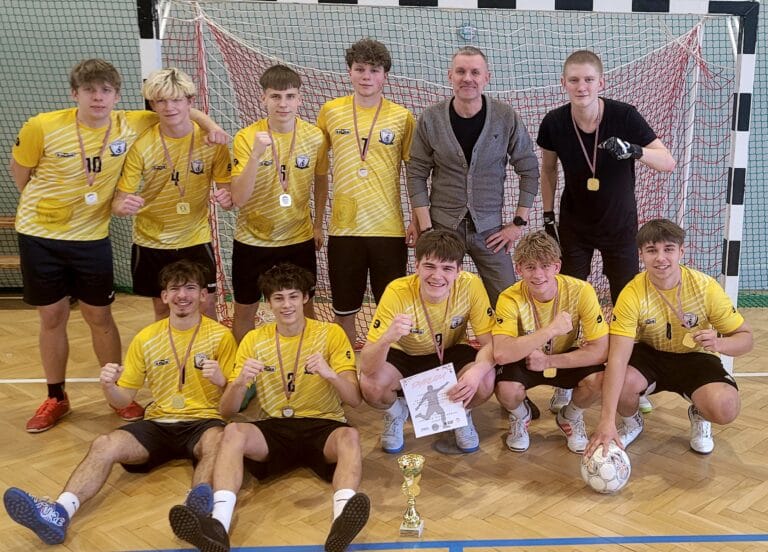 Zdjęcie grupowe męskiej drużyny sportowej w żółto-czarnych strojach, pozującej przed bramką na hali sportowej wraz z trenerem.