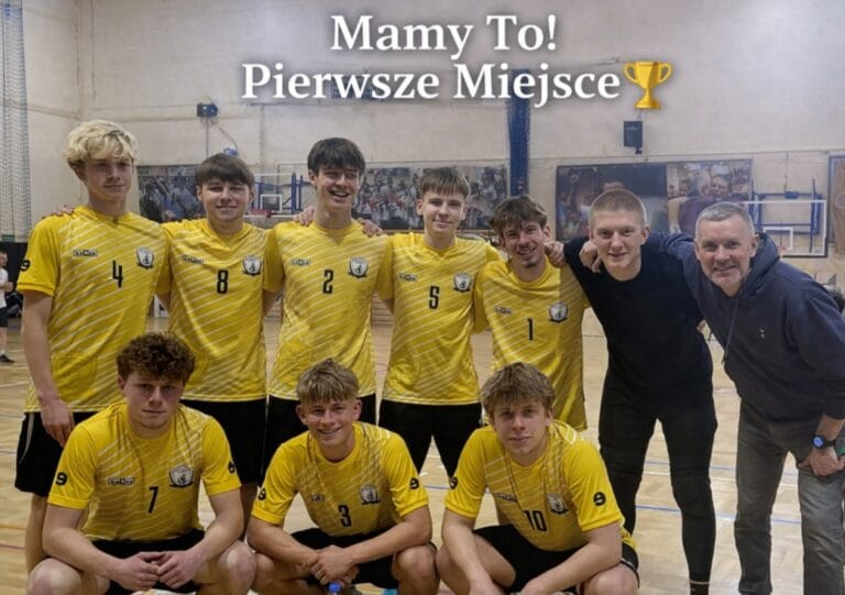 Zdjęcie drużyny sportowej pozującej na hali. Osoby mają żółte stroje z numerami i stoją oraz klęczą w dwóch rzędach. W tle widać siatkę oraz fragmenty sportowych banerów. U góry fotografii znajduje się napis: ‘Mamy To! Pierwsze Miejsce’ wraz z ikoną pucharu