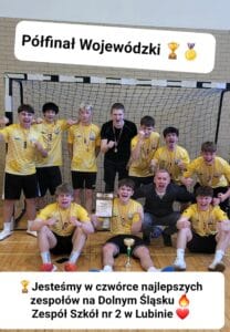 Pamiątkowe zdjęcie drużyny chłopców z Zespołu Szkół nr 2 w Lubinie, świętujących sukces w turnieju sportowym. Grupa nastolatków w żółto-białych koszulkach z medalami na szyjach pozuje z dyplomem i pucharem na tle bramki do piłki ręcznej. Wszyscy są bardzo radosni i krzyczą z entuzjazmu.