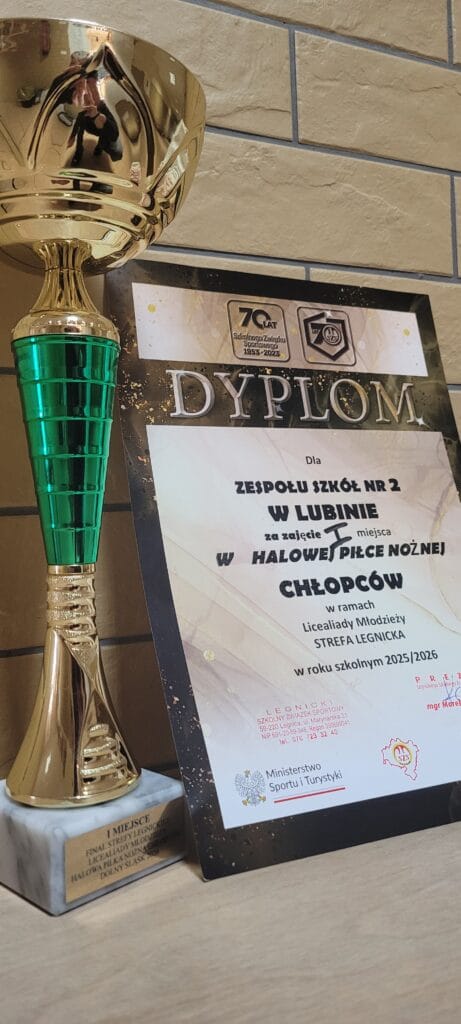 Złoty puchar i dyplom za zajęcie I miejsca w turnieju halowej piłki nożnej chłopców. Puchar ma zielone elementy dekoracyjne i marmurową podstawę z grawerowaną tabliczką. Obok stoi dyplom przyznany Zespołowi Szkół nr 2 w Lubinie w ramach Licealiady Młodzieży – Strefa Legnicka