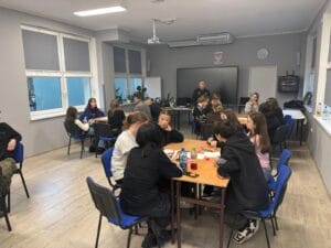 Osoby pracują przy stołach podczas zajęć w sali szkolnej, a jedna osoba stoi przy tablicy multimedialnej na końcu sali