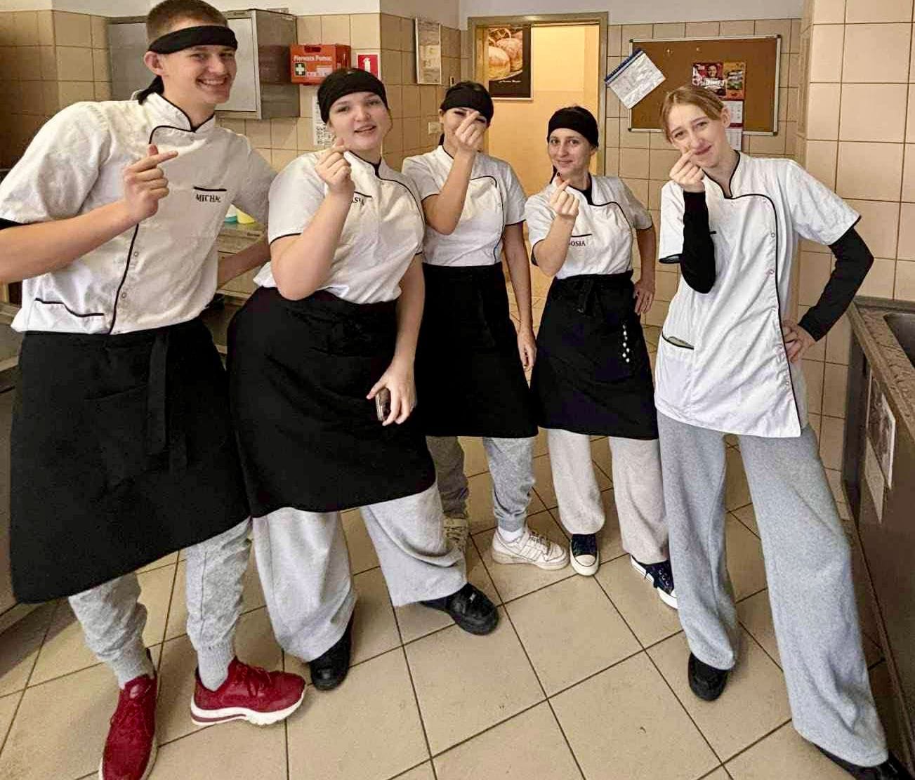 Grupa pięciorga uśmiechniętych młodych kucharzy pozuje w kuchni szkolnej lub warsztatowej. Wszyscy mają na sobie białe kitle z krótkim rękawem, czarne zapaski oraz czarne bandany lub czepki na głowach. Uczestnicy wykonują dłońmi popularny gest „koreańskiego serca” (skrzyżowane kciuki i palce wskazujące). Na kitle chłopaka po lewej stronie widoczne jest imię „MICHAŁ”, a u dziewczyny obok niego fragment imienia „KASIA”. W tle widać kafelkowane ściany, tablicę korkową oraz apteczkę pierwszej pomocy