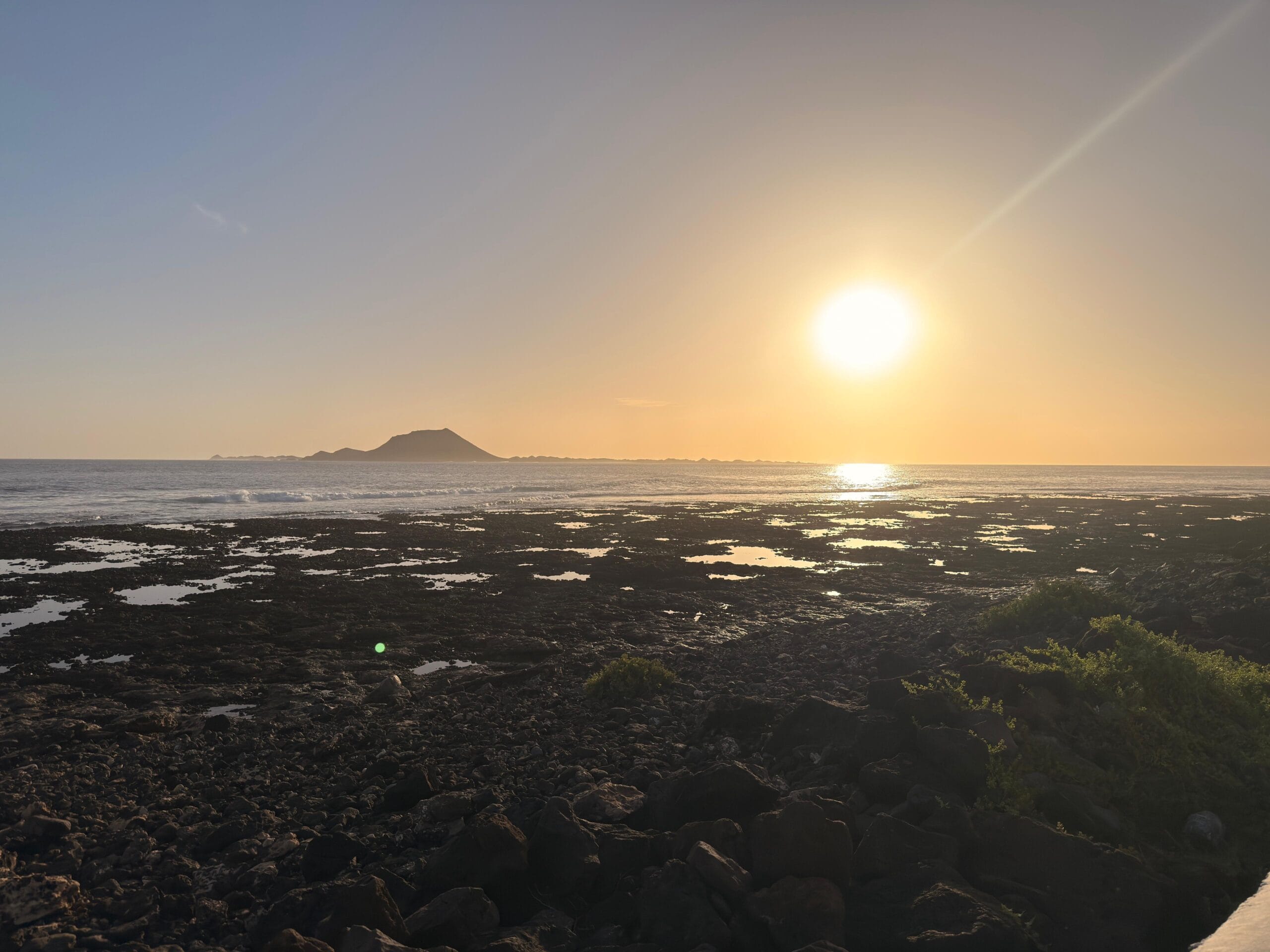 Szeroka panorama kamienistego wybrzeża oceanu podczas zachodu słońca. Słońce znajduje się nisko nad horyzontem, tworząc jasną smugę światła na wodzie. W oddali widać zarys górzystej wyspy, a na pierwszym planie ciemne skały i kępy niskiej roślinności