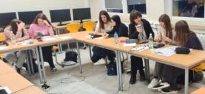 Grupa osób siedzi przy dwóch ustawionych w kształt litery L stołach w pracowni komputerowej i wykonuje zadania z użyciem terminali płatniczych. Na blatach znajdują się notatki, butelki z napojami, telefony oraz akcesoria do urządzeń. W tle widać rząd komputerów oraz okna