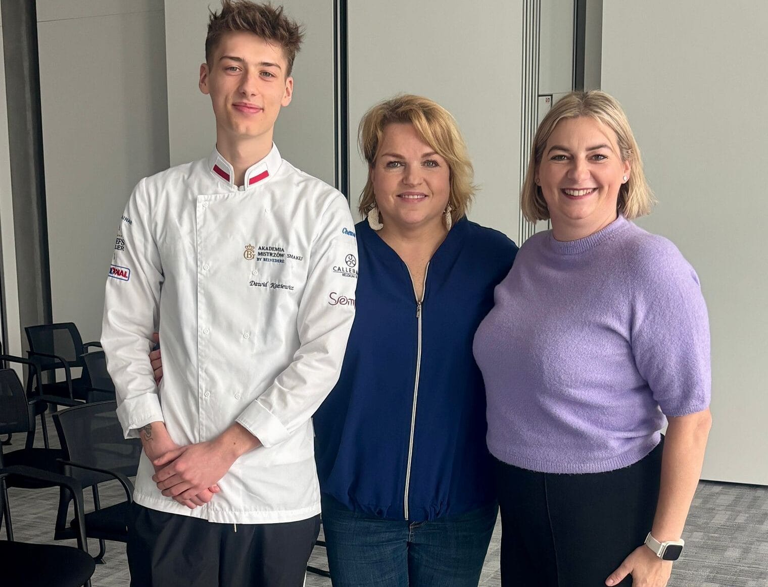 Zdjęcie przedstawia troje uśmiechniętych osób – młodego mężczyznę oraz dwie kobiety – pozujących wspólnie w nowoczesnym, jasnym pomieszczeniu. Po lewej stronie stoi mężczyzna w białym kitlu kucharskim z logo „Akademia Mistrzów Smaku by Belvedere” oraz wyhaftowanym nazwiskiem Dawid Kasiewicz na piersi; na rękawach jego munduru widoczne są mniejsze logotypy partnerów, m.in. Rational i Callebaut. W środku stoi kobieta w ciemnoniebieskiej rozpinanej bluzie i jeansach, a po prawej kobieta w liliowym swetrze z krótkim rękawem i czarnych spodniach, mająca na ręku biały smartwatch. Grupa znajduje się na tle szarych ścian i przeszklonych drzwi, a po lewej stronie w głębi widoczne są rzędy czarnych krzeseł biurowych na wzorzystej, szarej wykładzinie.