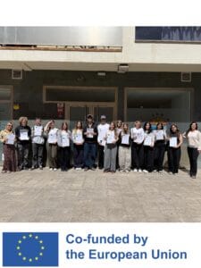 Grupa 16 osób stoi w rzędzie przed nowoczesnym budynkiem. Większość osób trzyma w rękach białe dyplomy lub certyfikaty z logo Unii Europejskiej. Poniżej zdjęcia znajduje się logotyp „Co-funded by the European Union”