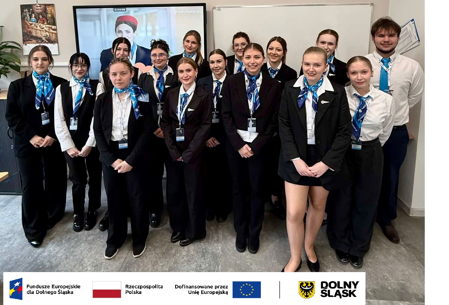 Pozowane zdjęcie grupowe czternastu uczestniczek i jednego uczestnika kursu personelu pokładowego w sali wykładowej. Wszyscy są ubrani w ciemne mundury z niebieskimi apaszkami. Poniżej zdjęcia znajduje się pasek z logotypami Funduszy Europejskich dla Dolnego Śląska, Rzeczypospolitej Polskiej, Unii Europejskiej oraz Województwa Dolnośląskiego.