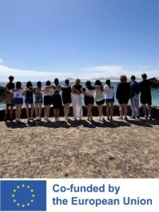 Trzy młode osoby siedzą na piaszczystej wydmie tyłem do obiektywu, patrząc w stronę oceanu i gór w oddali. Pod zdjęciem znajduje się biały pasek z logotypem Unii Europejskiej oraz napisem „Co-funded by the European Union”.