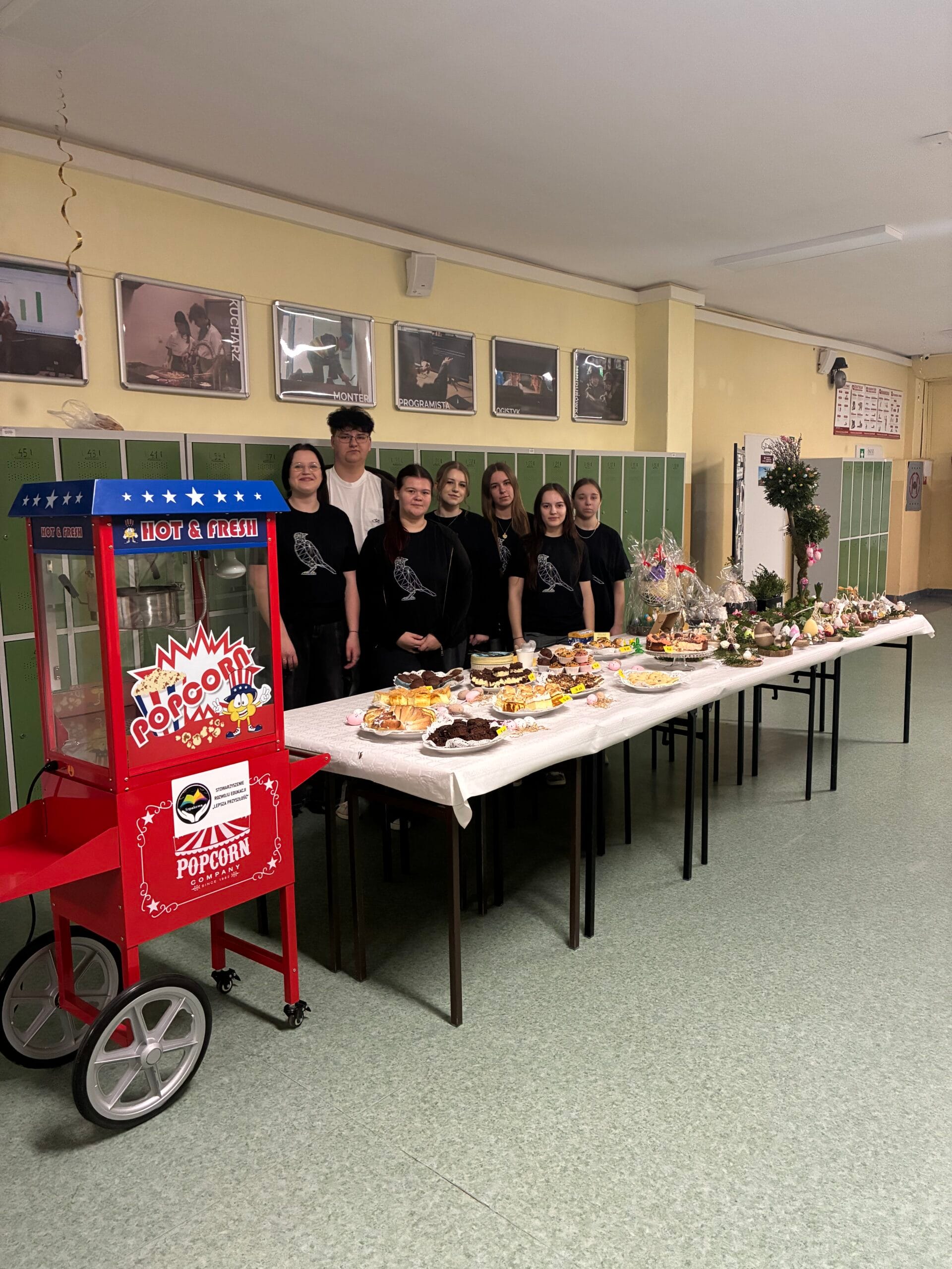 Grupa młodzieży stoi za długim stołem z wielkanocnymi wypiekami i ozdobami na szkolnym korytarzu. Obok stołu po lewej stronie znajduje się czerwona maszyna do popcornu w stylu retro. W tle widoczne są zielone szafki i zdjęcia w ramkach na ścianie.