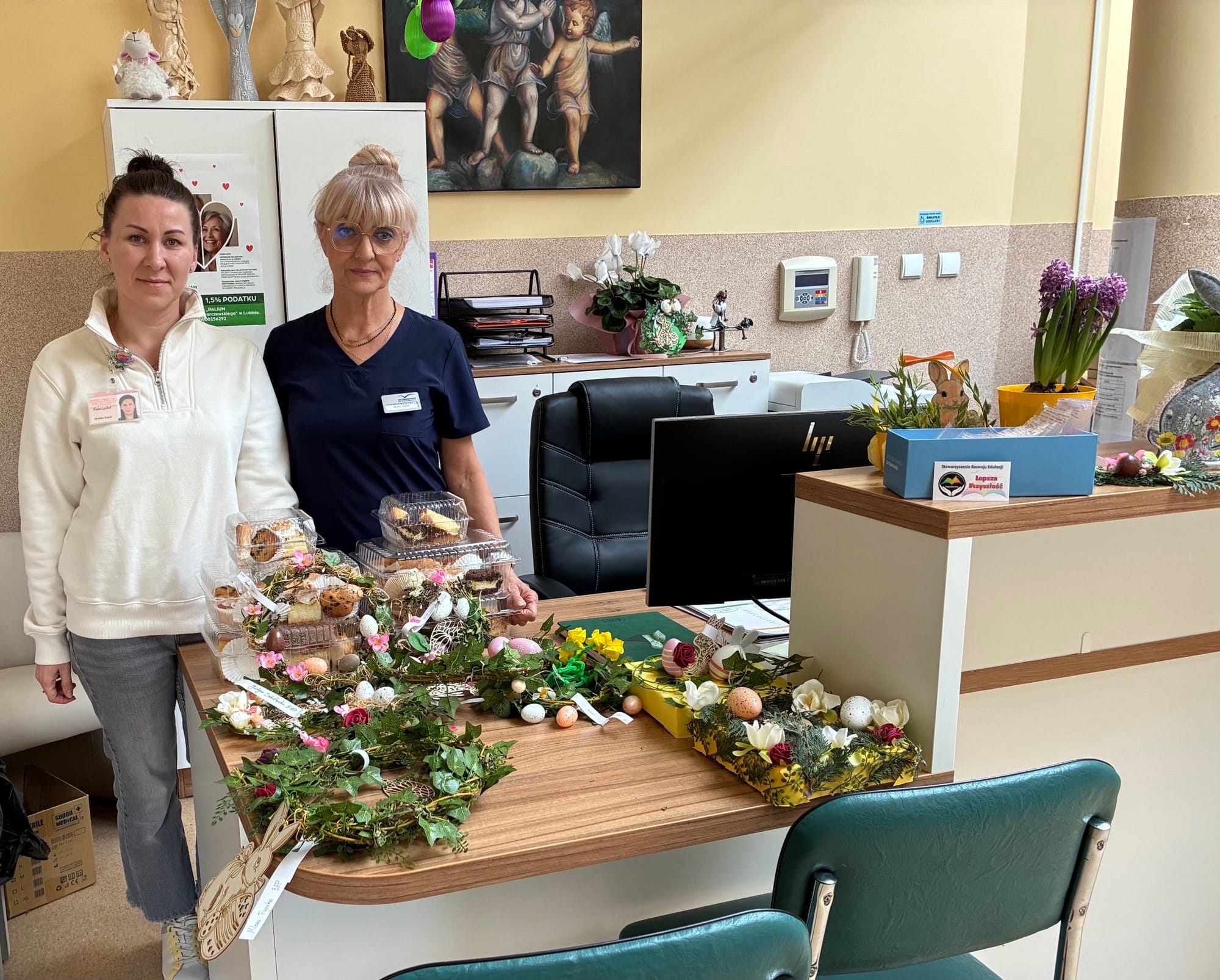 Dwie kobiety w strojach medycznych stoją za biurkiem w gabinecie lub rejestracji, na którym ułożono liczne stroiki wielkanocne oraz ciasta w plastikowych pojemnikach. W tle widoczne są kwiaty doniczkowe oraz obraz przedstawiający anioły.