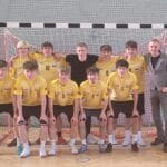 Oficjalne zdjęcie grupowe męskiej drużyny futsalu Zespołu Szkół nr 2 w Lubinie wraz z trenerem, pozujących w hali sportowej na tle bramki po zdobyciu tytułu Mistrza Dolnego Śląska. Zawodnicy są w żółto-białych strojach, a na szyjach mają medale.
