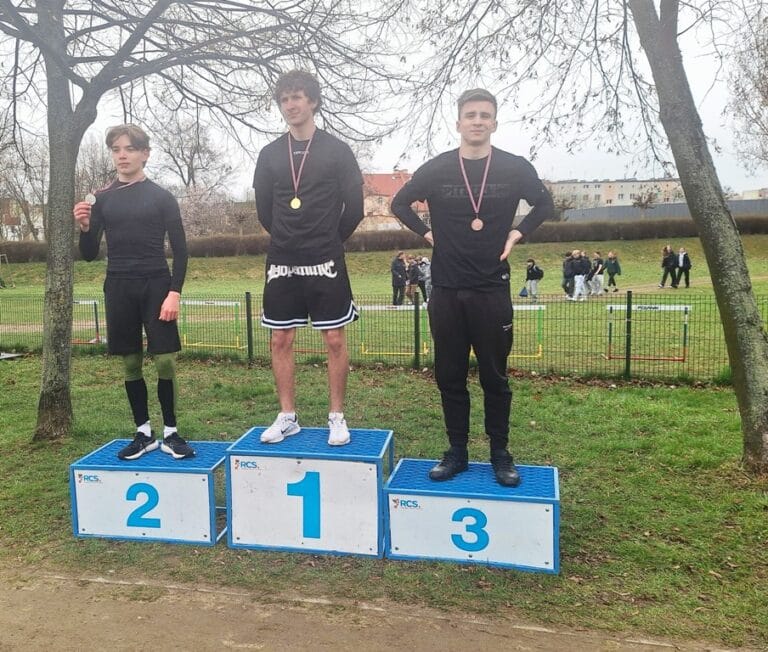 Trzech młodych sportowców na podium podczas zawodów lekkoatletycznych na świeżym powietrzu. Na najwyższym stopniu z numerem 1 stoi zwycięzca w złotym medalu, po jego lewej stronie na stopniu z numerem 2 stoi srebrny medalista, a po prawej na stopniu z numerem 3 – brązowy medalista. Wszyscy są ubrani w stroje sportowe. W tle widoczna jest trawiasta murawa, drzewa bez liści, płot oraz inni uczestnicy wydarzenia.