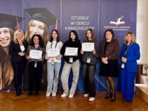 Grupa dziewięciu osób stoi przed ścianką z napisami „Studiuj w sercu Karkonoszy” oraz „Karkonoska Akademia Nauk Stosowanych”. Cztery osoby trzymają certyfikaty. Wszystkie osoby mają identyfikatory na smyczach. Zdjęcie wykonane w sali, na tle ścianki promocyjnej