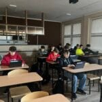 Grupa uczniów siedzi w ławkach w klasie i pracuje na laptopach. Wszyscy są skupieni na ekranach swoich urządzeń. Sala jest oświetlona światłem dziennym wpadającym przez duże okna po prawej stronie. W głębi pomieszczenia widoczna jest zabudowa przypominająca barek lub aneks kuchenny.