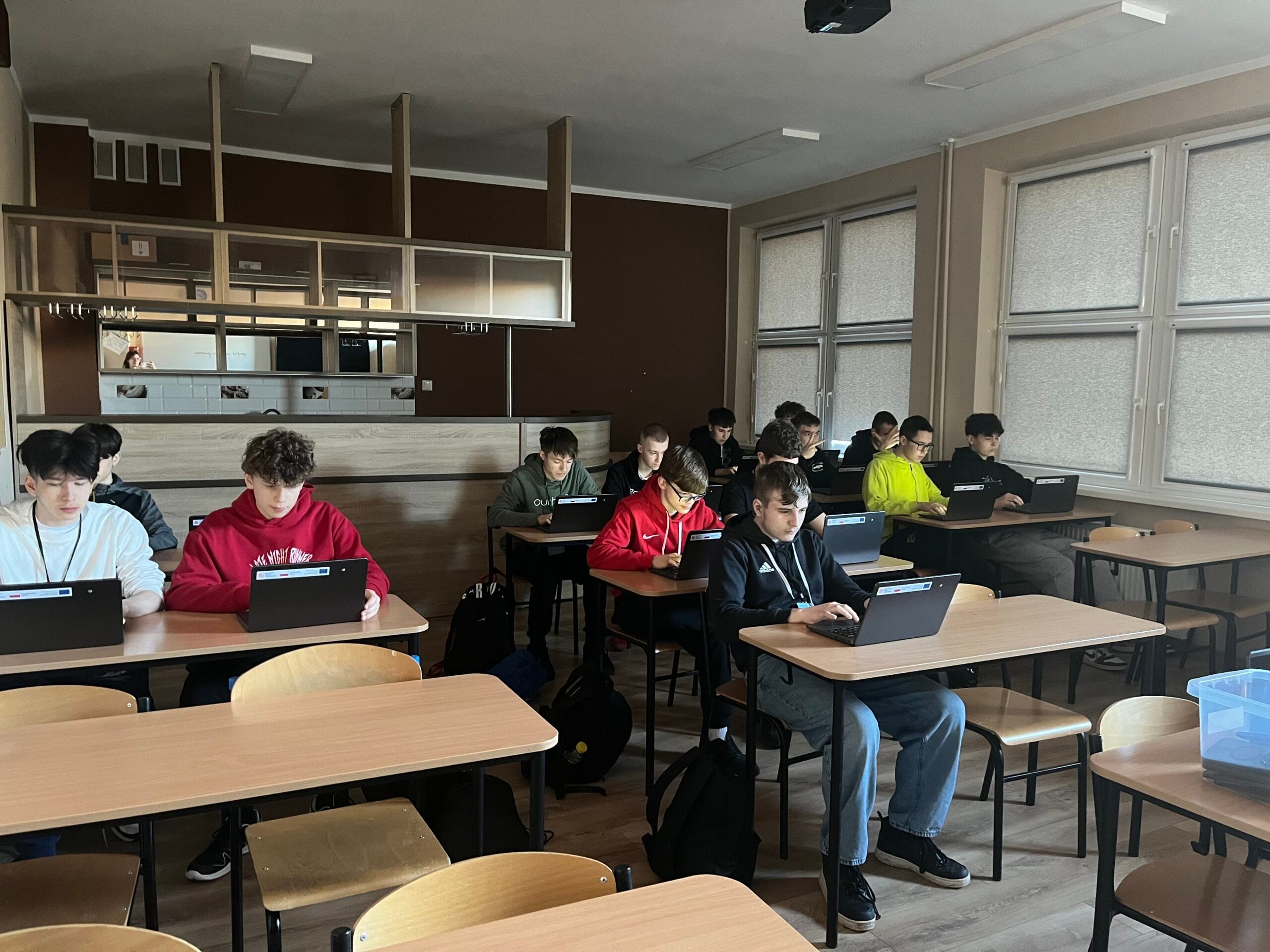 Grupa uczniów siedzi w ławkach w klasie i pracuje na laptopach. Wszyscy są skupieni na ekranach swoich urządzeń. Sala jest oświetlona światłem dziennym wpadającym przez duże okna po prawej stronie. W głębi pomieszczenia widoczna jest zabudowa przypominająca barek lub aneks kuchenny.