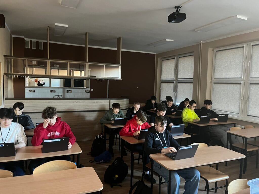 Grupa uczniów siedzi w ławkach w klasie i pracuje na laptopach. Wszyscy są skupieni na ekranach swoich urządzeń. Sala jest oświetlona światłem dziennym wpadającym przez duże okna po prawej stronie. W głębi pomieszczenia widoczna jest zabudowa przypominająca barek lub aneks kuchenny.