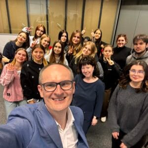Zdjęcie typu selfie przedstawiające grupę uśmiechniętej młodzieży z dwojgiem opiekunów. Na pierwszym planie uśmiechnięty mężczyzna w okularach i niebieskiej marynarce trzyma aparat. Za nim stoi grupa kilkunastu osób; niektórzy wykonują gesty zwycięstwa (V) lub kciuki w górę. Wszyscy znajdują się w pomieszczeniu o nowoczesnym wystroju.