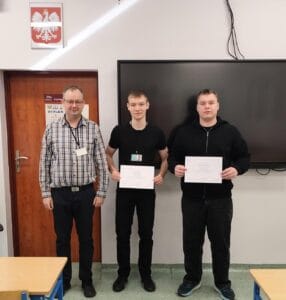 Trzech mężczyzn stoi w sali lekcyjnej na tle białej ściany i czarnej tablicy multimedialnej. Dwaj młodsi mężczyźni po prawej trzymają białe dyplomy. Mężczyzna po lewej, w okularach i kraciastej koszuli, stoi obok nich. Nad drzwiami po lewej stronie wisi godło Polski.