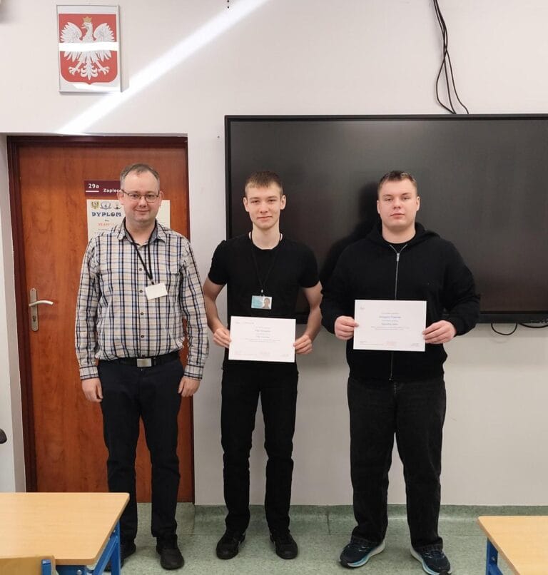 Trzech mężczyzn stoi w sali lekcyjnej na tle białej ściany i czarnej tablicy multimedialnej. Dwaj młodsi mężczyźni po prawej trzymają białe dyplomy. Mężczyzna po lewej, w okularach i kraciastej koszuli, stoi obok nich. Nad drzwiami po lewej stronie wisi godło Polski.