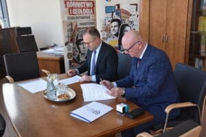 Dwie osoby siedzą przy stole w biurze i podpisują dokumenty. Na stole znajdują się teczki z dokumentami, długopisy, szklanki z wodą oraz talerz z przekąskami. W tle widoczne są meble biurowe oraz baner z napisem „Górnictwo i Geologia – Studia inżynierskie i magisterskie