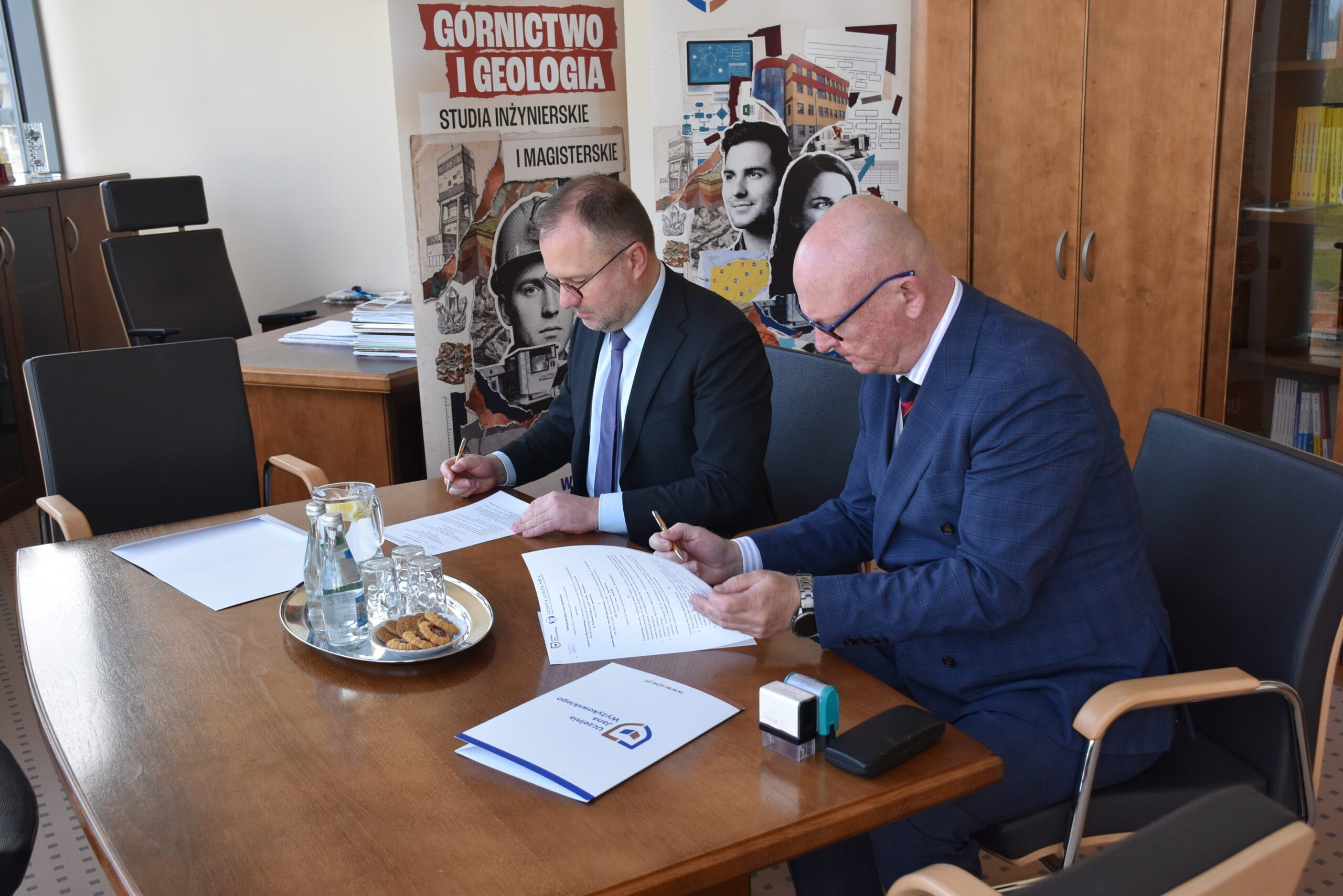 Dwie osoby siedzą przy stole w biurze i podpisują dokumenty. Na stole znajdują się teczki z dokumentami, długopisy, szklanki z wodą oraz talerz z przekąskami. W tle widoczne są meble biurowe oraz baner z napisem „Górnictwo i Geologia – Studia inżynierskie i magisterskie