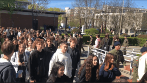 Tłum uczniów Zespołu Szkół nr 2 w Lubinie czekający przed wejściem do budynku. Wśród cywilnej młodzieży widoczni są uczniowie klas mundurowych. W tle widać bloki mieszkalne i drzewa w wiosennej aurze.