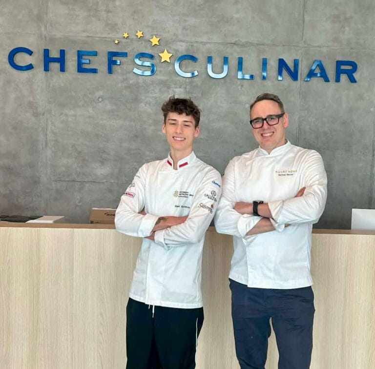 Dwóch uśmiechniętych kucharzy w białych kitlach stoi obok siebie z założonymi rękami na tle szarej ściany z dużym niebieskim napisem „CHEFS CULINAR” i złotymi gwiazdkami.
