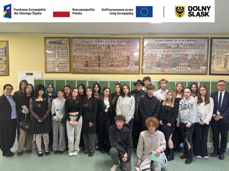 Zdjęcie grupowe młodzieży i dorosłych w szkolnym korytarzu na tle zielonych szafek i tablic pamiątkowych Zespołu Szkół nr 2 w Lubinie. W centrum grupy stoi mężczyzna w czarnym stroju kucharskim i kapeluszu. U góry grafiki widnieje biały pas z logotypami: Fundusze Europejskie dla Dolnego Śląska, flaga Polski z napisem Rzeczpospolita Polska, flaga Unii Europejskiej oraz herb województwa dolnośląskiego.