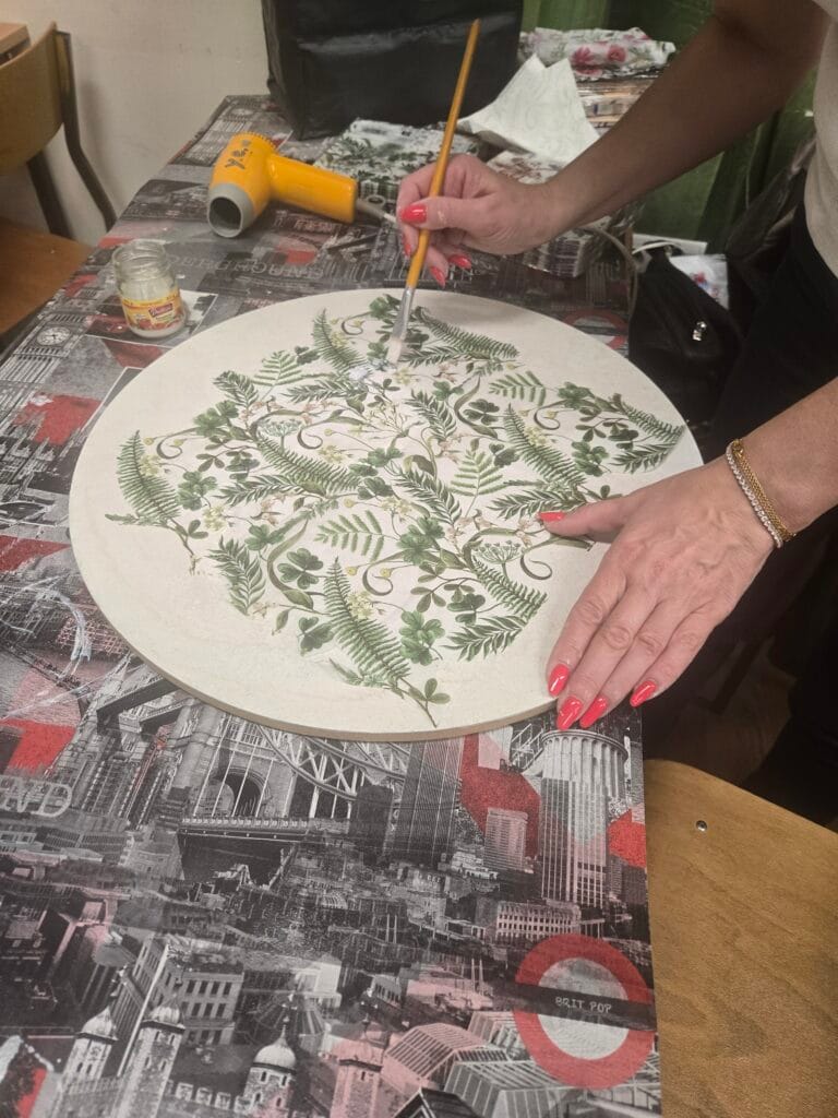 Zbliżenie na dłonie kobiety z różowym manikiurem, która nakłada pędzlem klej na okrągły, jasny blat. Na środku blatu rozłożona jest serwetka z gęstym motywem zielonych liści paproci i koniczyny. Praca odbywa się na stole zabezpieczonym podkładką z czarno-biało-czerwonym motywem Londynu. W tle widoczna jest mała żółta suszarka oraz słoiczek z klejem