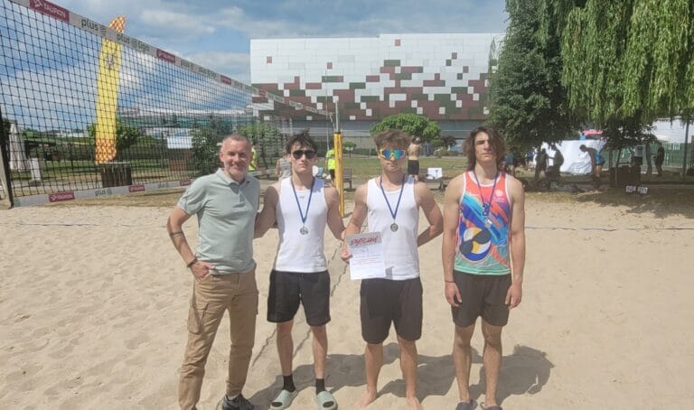 Pamiątkowe zdjęcie na piaszczystym boisku do siatkówki plażowej. Od lewej stoi mężczyzna w jasnej koszulce polo i spodniach bojówkach, a obok niego trzech młodych zawodników z medalami na szyjach. Dwóch z nich ma na sobie białe tank topy, a trzeci kolorową koszulkę sportową. Środkowy zawodnik trzyma biały dyplom z czerwonym napisem 'DYPLOM'. Po lewej stronie kadru widoczna jest siatka z logo sponsorów (Plus, Tauron), a w tle nowoczesny budynek z charakterystyczną mozaiką oraz drzewa