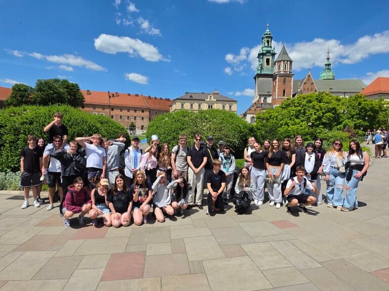 Duża grupa młodzieży pozuje do wspólnego zdjęcia na dziedzińcu Zamku Królewskiego na Wawelu. W tle, pod bezchmurnym niebem, widać wieże Katedry Wawelskiej oraz zielone krzewy