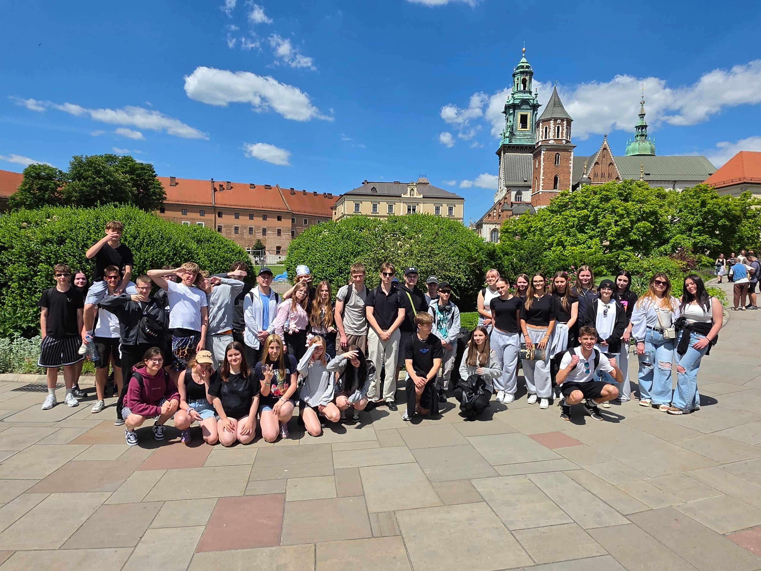 Duża grupa młodzieży pozuje do wspólnego zdjęcia na dziedzińcu Zamku Królewskiego na Wawelu. W tle, pod bezchmurnym niebem, widać wieże Katedry Wawelskiej oraz zielone krzewy