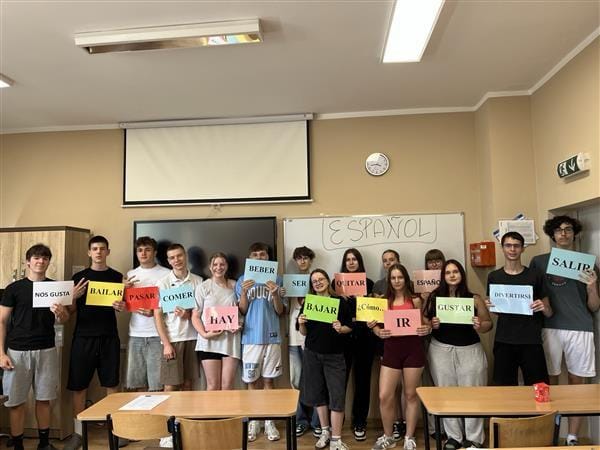 Grupa młodzieży stoi w klasie lekcyjnej i trzyma kolorowe kartki z hiszpańskimi słowami. W tle widać białą tablicę z napisem ESPAÑOL