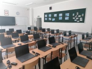 Nowoczesna szkolna pracownia komputerowa z rzędami biurek, na których stoją otwarte czarne laptopy. Na ścianie w głębi widać zieloną tablicę informacyjną oraz godło Polski.
